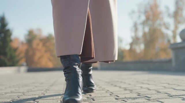 Coatclad woman walking on cobblestone pavement, slow confident stride, black leather boots with block heel, soft golden sunlight, crisp autumn trees and park balustrade, textured paving, fashion