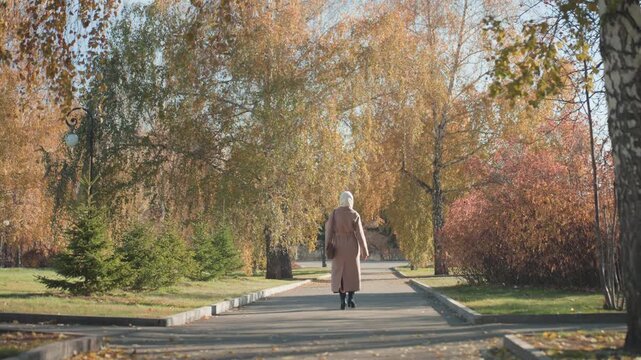 White senior woman brisk autumn walk along park walkway, determined stride down sunlit path, coat and bag, fallen leaves underfoot, crisp midday light, solitary commuter mood with calm focus