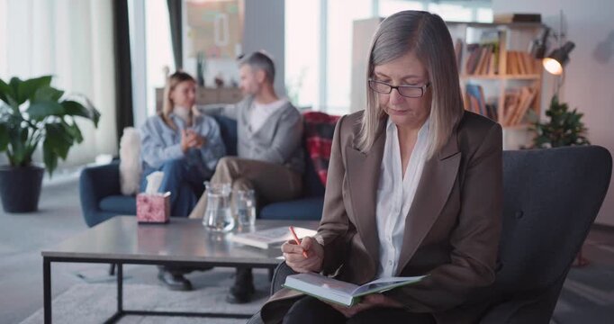 Indoor portrait of caucasian family therapist mature professional woman posing to camera. Couple married man with woman talking in background.