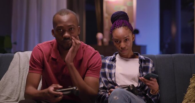 African american love couple at home. Excited male using console gamepad playing online virtual game while his girlfriend using mobile phone.