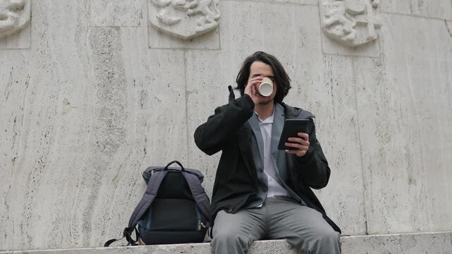 Young student reading an ebook and drinking coffee while sitting on stone steps in amsterdam with his backpack