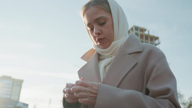 Young woman outdoors adjusting scarf, city morning scene with wool coat and headscarf, construction site in background, soft sunlight and pale sky, contemplative expression with hands fidgeting, slow