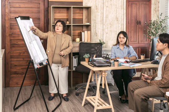 Senior Asian female manager leads a business presentation, pointing to a chart on a flipboard. An experienced team leader trains her junior colleagues in a modern office.