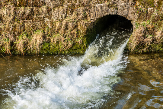 Water flows from a stone drain into a river in a rural area during daylight hours near a natural landscape with grass and rocks