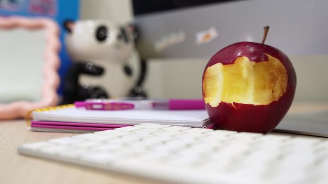 A cinematic close-up of a bitten red apple on a modern office desk next to a keyboard and notebook. Perfect for productivity, healthy lifestyle, and workspace-themed content.