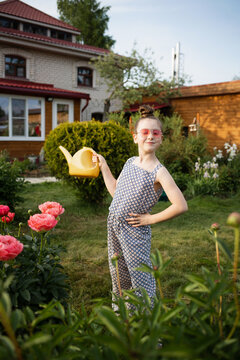child fashionable girl with watering can, stands in backyard among flowers and flower beds. summer vacation in countryside, vacations