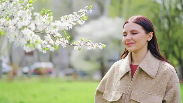 Young woman smiles brightly, finding joy and serenity amidst beautiful spring blossoms in sunlit park. Fresh air and vibrant nature evoke feelings of peace.