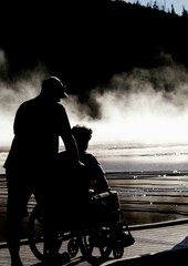Silhouette of a man assisting a person in a wheelchair by a misty lake captures emotional support and inclusive travel opportunities. © Simeon