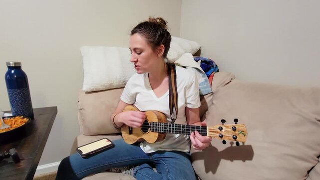 Woman sings while playing ukulele on a couch indoors, performing music at home with a casual acoustic setup, smartphone lyrics nearby, and a relaxed intimate lifestyle scene.