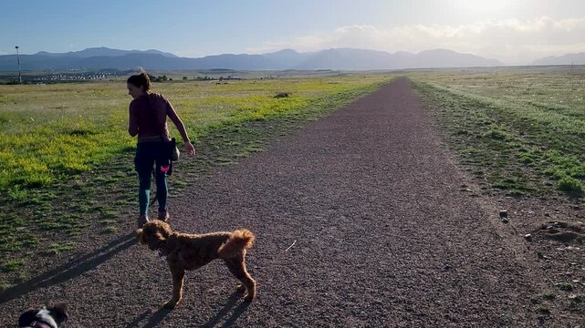 Woman gives her dog a treat on a wide gravel trail through open grassland, with distant mountains and warm evening light creating a peaceful outdoor lifestyle and pet bonding scene.