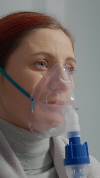 Vertical Screen: A woman relaxes on a couch while wearing a mask connected to a nebulizer. The device releases steam to help treat her cough and respiratory issues.