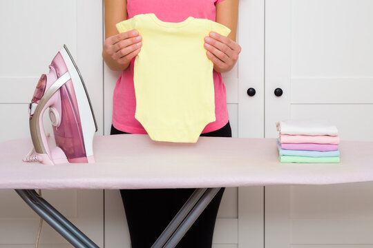 Young adult mother hands holding clean ironed yellow newborn bodysuit at white wardrobe at home room. Closeup. Front view.