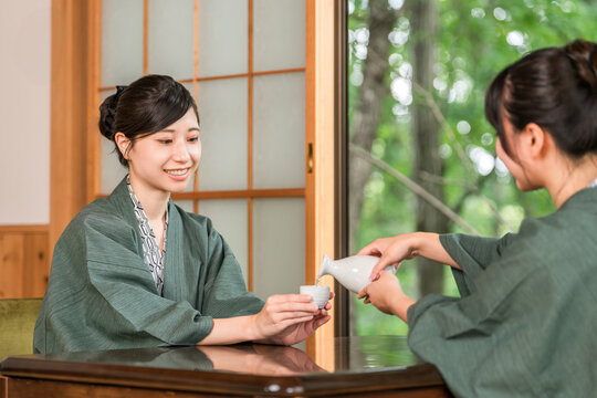 A woman and her friend drinking sake on the veranda of a Japanese-style room in a traditional Japanese inn, both wearing yukata robes.