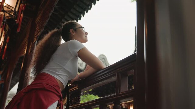 Young woman gazing from temple, low angle view under ornate eaves with wooden beams and carved railing, red skirt and ponytail, glasses, bright skyline beyond courtyard, curious traveler watching