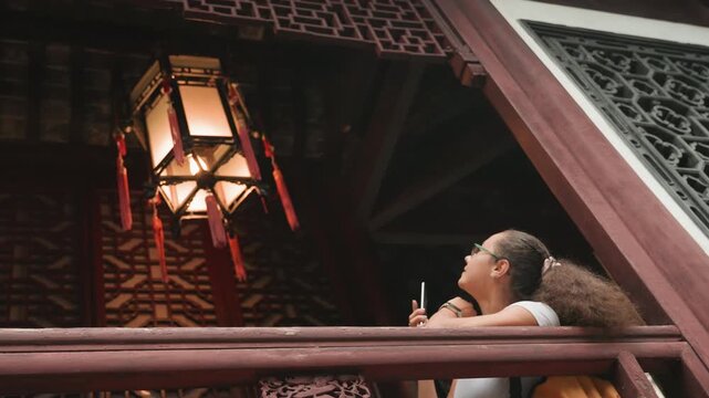 Young black woman admiring hanging lantern, warm glow illuminating carved eaves and tassels, smartphone pointed upward, contemplative gaze, wooden ceiling and ornate lattice backdrop, dusk atmosphere,