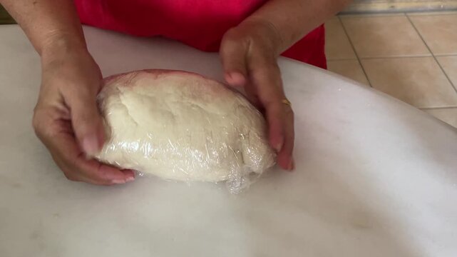 A close-up shot captures hands carefully wrapping a smooth white dough with transparent plastic wrap on a white table to let it rest or proof for traditional handmade buns.