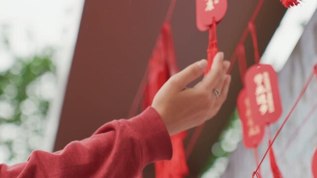 Closeup red tassel wish tags hanging, gentle hand in red sleeve reaches up to touch wooden tags and braided tassels, soft bokeh background, delicate ring on finger, calm ritual atmosphere at outdoor