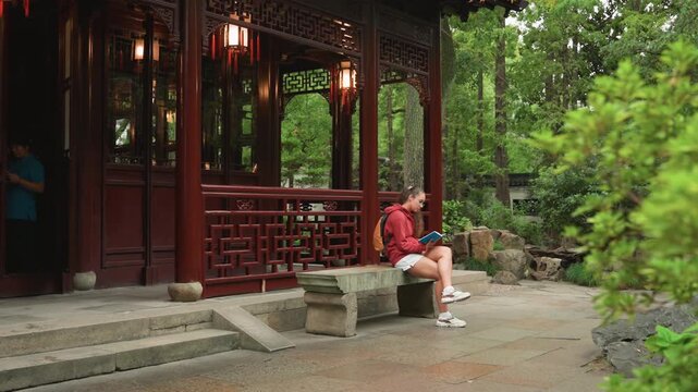 Caucasian woman sitting red pavilion stone steps green foliage checking phone contemplative pause light rain asian garden architecture tourist backpack casual clothing peaceful moment
