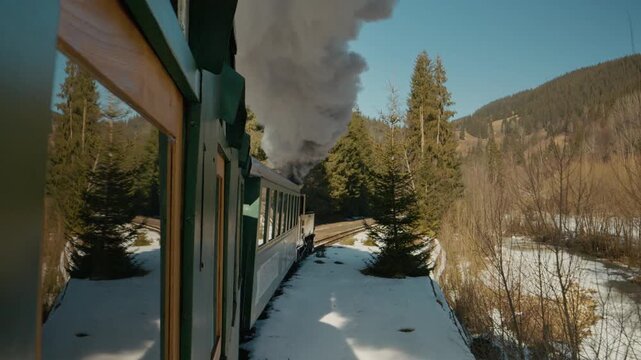 A steam train moves through a winter forest. Snow covers the ground as trees line the tracks. The locomotive puffs steam into the cold air.