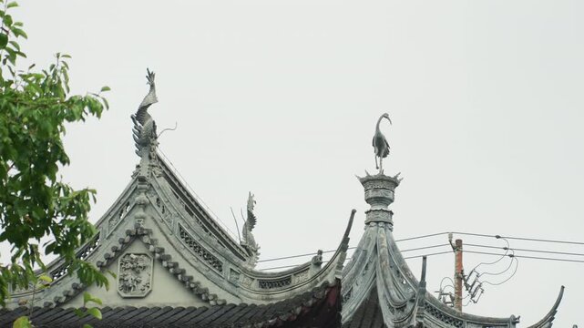 Ornate temple roof under overcast sky, detailed eaves and guardian statues framed by leafy tree, weathered carvings and cornice lines evoke historic craftsmanship and serene atmosphere