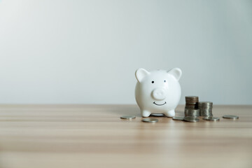 White piggy bank smiles softly on wooden surface. Coins scattered nearby hint at savings and...