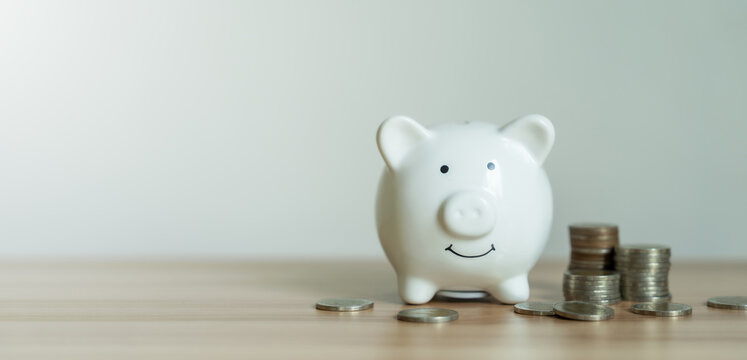 White piggy bank smiles gently on wooden surface. Stacks of coins lie beside it, suggesting savings. Soft lighting highlights its cheerful expression. Ideal for budgeting, finance