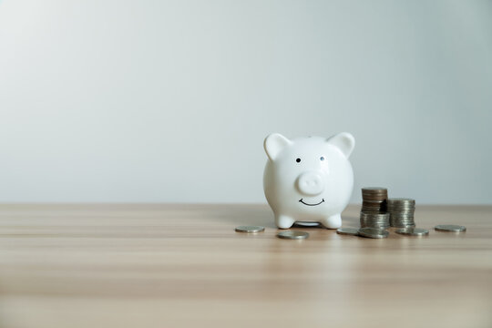 White piggy bank smiles softly on wooden surface. Coins scattered nearby hint at savings and growth. Minimalist background lets focus remain on financial theme. Ideal for budgeting, finance