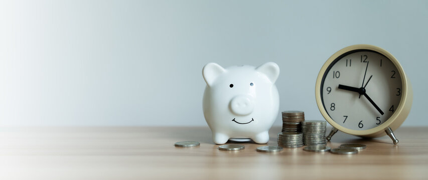 Minimalist scene with piggy bank and clock on wooden surface. Coins stacked beside the piggy bank. Time and savings shown in simple, clean composition. Ideal for financial, budgeting
