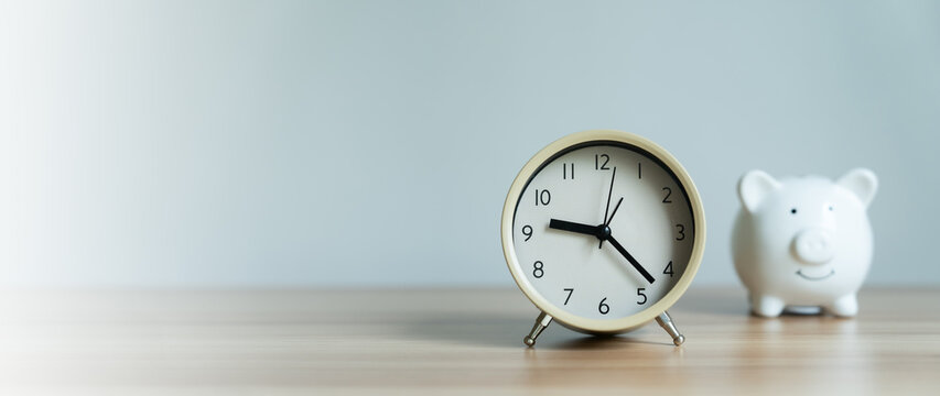 Simple clock sits on wooden desk. White piggy bank rests beside it. Soft light fills calm, minimal background. No people, no motion, no noise. Perfect for budgeting, time management, savings concepts