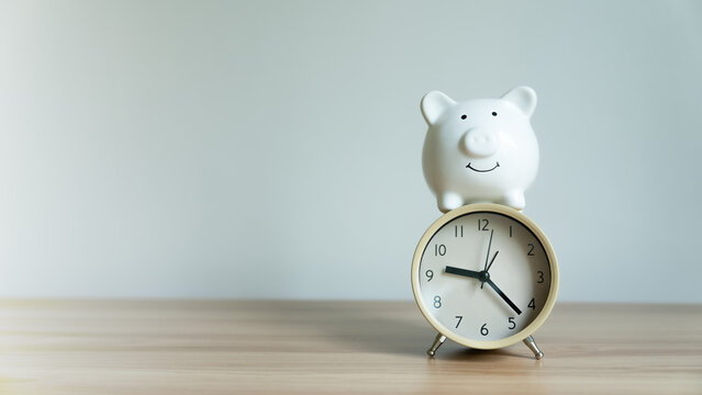 White piggy bank smiles atop round alarm clock. Clock shows time, suggesting urgency or deadlines. Simplicity of wood and light wall creates calm focus. Ideal for finance, budgeting