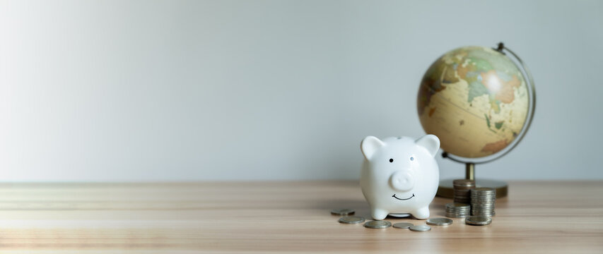 White piggy bank sits beside globe and coins. Wooden surface holds all items neatly. Globe suggests global financial awareness. Coins represent savings or investment growth
