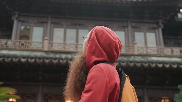 Hooded traveler at historic temple, young woman backpacker and architecture student, pauses to gaze upward at ornate wooden eaves and carved facade, soft lantern light, overcast sky, curious