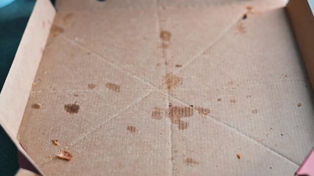 A hand reaches for the last slice of pizza in an empty box. The box shows sauce stains and a few toppings scattered inside, indicating a recent meal