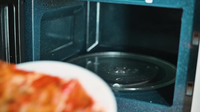 A hand reaches into a microwave to place a slice of leftover pizza on a plate. The pizza is then removed after heating, showing a quick and simple meal preparation at home