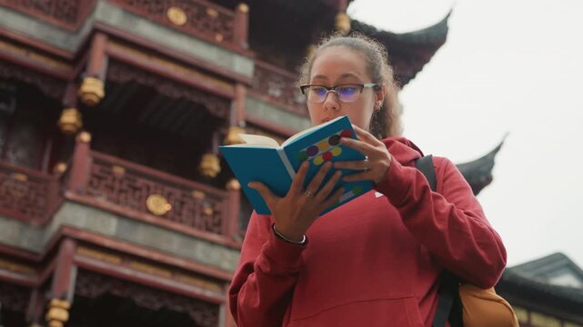 Young woman reading guidebook at temple, blue notebook held close, red hoodie and backpack, ornate pagoda with carved eaves in background, calm introspective expression, travel blogger style shot,