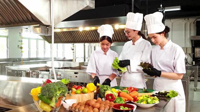 Three chefs in a commercial kitchen prepare fresh vegetables, wearing white uniforms and gloves, surrounded by colorful produce and eggs during a culinary training session.