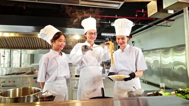 Three chefs in a professional stainless-steel kitchen prepare steaming pasta, smiling as one lifts noodles with tongs while another plates the dish during a culinary training session.