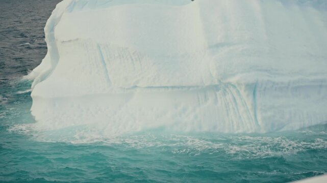 Ship passes extremely close to a massive striated Antarctic iceberg wall, its textured blue-white surface rising dramatically above choppy turquoise polar waters