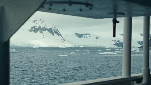 Cinematic POV shot framed through an open expedition ship hatch looking out across ice-dotted polar waters toward jagged snow-covered Antarctic mountains under overcast skies