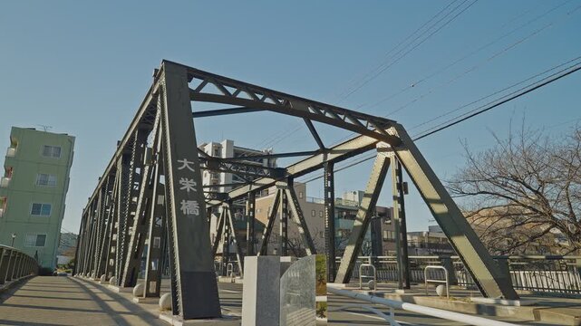 Dark steel truss bridge with Japanese nameplate stands against vivid blue sky in Tokyo, with urban buildings and bare trees alongside, captured as a static low-angle shot from the bridge deck.