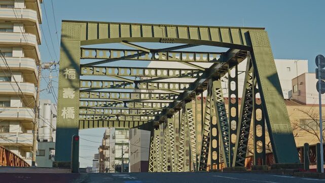 Fukuju Bridge's green steel truss forms a bold gateway arch over a quiet urban street in Tokyo, with Japanese nameplate and residential buildings alongside, captured as a static straight-on wide shot.