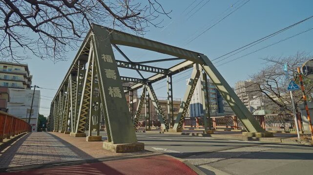 Fukuju Bridge's green steel truss stands at a busy urban intersection in Tokyo, with Japanese nameplate, sidewalk, bare winter trees, and city buildings visible, captured as a static wide ground-level