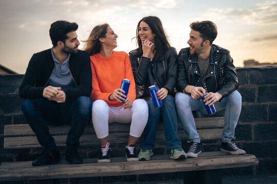 Group of young friends laughing and drinking energy drinks on a rooftop bench.