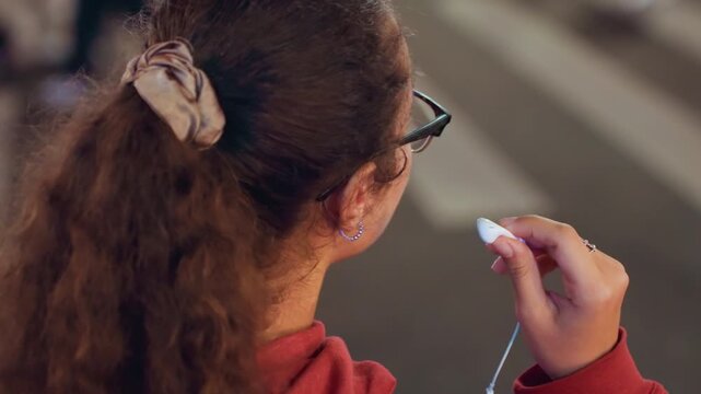 Young woman at crosswalk with earbuds under streetlights wearing glasses and scrunchie, adjusting earbud while looking both ways, cinematic nighttime city portrait conveying quiet contemplation