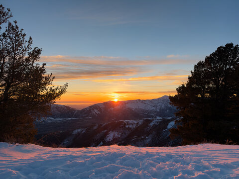Sunset Framed by Pine Trees Over Snowy Mountain Landscape