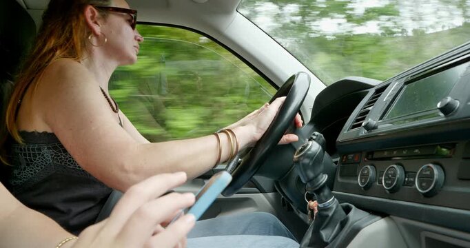 A woman is driving, and the hands of the passenger in the front seat, holding a phone, are guiding the way
