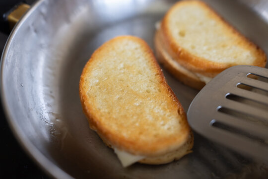 Golden toast buns sizzle in a pan. Cheese melts between bread slices. Spatula rests beside them, ready to flip. Pan shows signs of recent cooking. Food looks warm, crisp, and freshly made