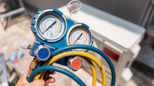 Technician's hand holding a manifold gauge with hoses in front of an outdoor air conditioning unit, close-up of a pressure gauge and hoses for refrigerant checking during AC maintenance