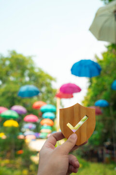Holding wooden shield with checkmark against colorful umbrellas background. Travel insurance, outdoor safety, and vacation protection concept. Secure leisure, verified tour, and fun holiday.