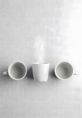 Fototapeta premium Three white coffee mugs on a table with steam rising from one mug in a minimalist still life photography style with a monochromatic color scheme and soft natural lighting